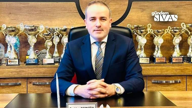 Bir masa üzerinde oturan bir kişi, arkasında kupa ve ödül trofeleri var.