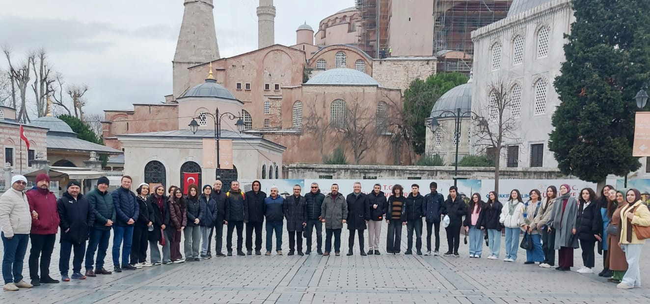 Tarihi kubbeli ve minareli bir yapının önünde, taş avluda sıralanmış kalabalık bir grup görülüyor.