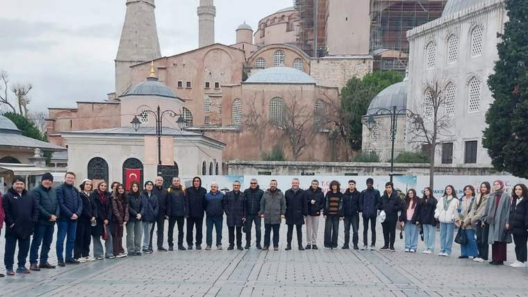 Tarihi kubbeli ve minareli bir yapının önünde, taş avluda sıralanmış kalabalık bir grup görülüyor.