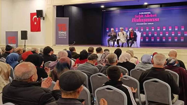 Salonda oturan kalabalık, sahnedeki konuşmacıları izliyor; arkadaki ekranda “Şehit Aileleri Buluşması” yazıyor.