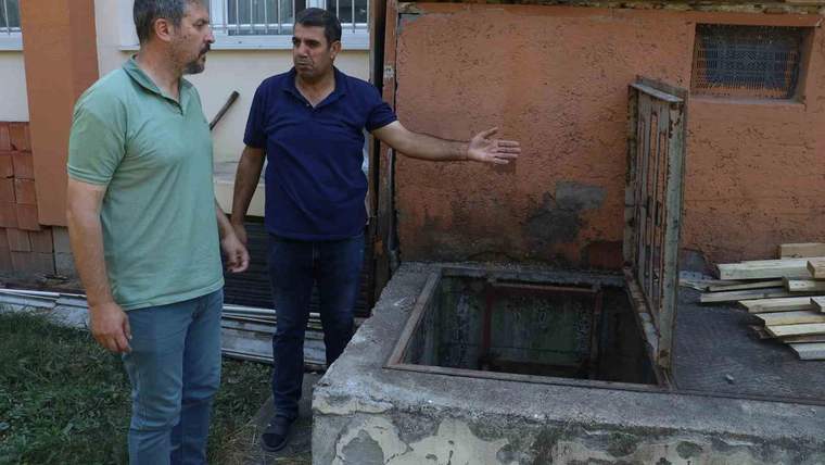 İki erkek, okul binasının yanında üzeri açık bir kuyunun başında duruyor; biri kuyuyu işaret ediyor.
