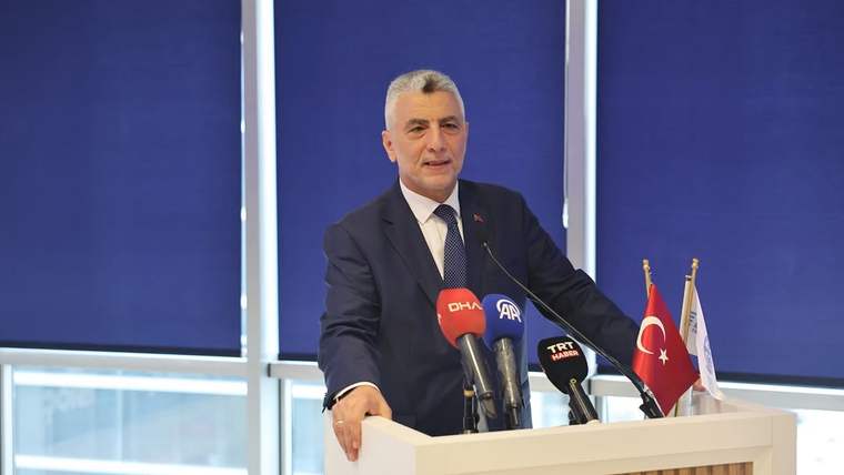 Kürsüde konuşan takım elbiseli bir erkek, önündeki mikrofonlar ve Türk bayrağının yanında duruyor.
