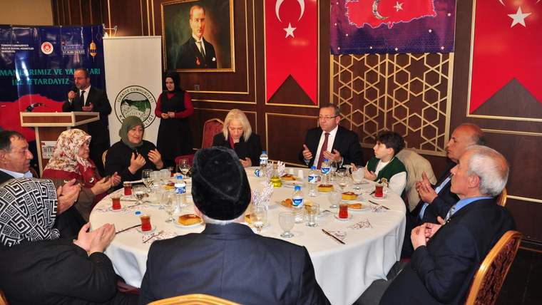 Türk bayrakları ve panoların bulunduğu salonda, yuvarlak masada yemek yiyen ve sohbet eden kişiler