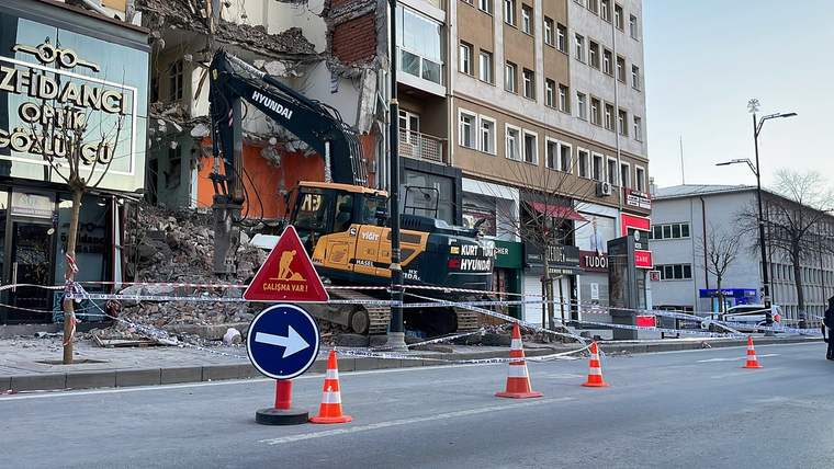 Bir inşaat makinesi, yıkım çalışması yapılan bir binanın önünde duruyor; çevresinde güvenlik şeritleri ve uyarı levhaları var.