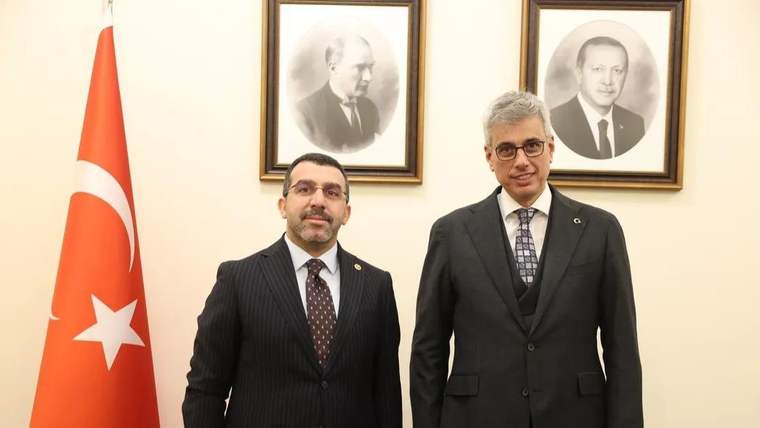 Türk bayrağı yanında takım elbiseli iki erkek, duvarda çerçeveli iki portre önünde duruyor.