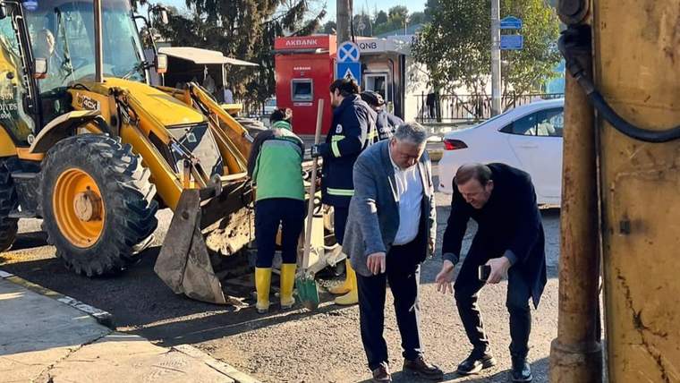 Sarı kazı makinesi yanında üç kişi, kaldırım kenarındaki kazı ve asfalt çalışması alanında duruyor.