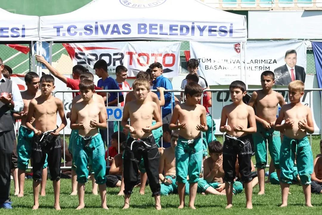 Yeşil ve siyah güreş kıyafetleri giyen çocuklar çim alanda sıralanıyor; arkada T.C. Sivas Belediyesi pankartı var.