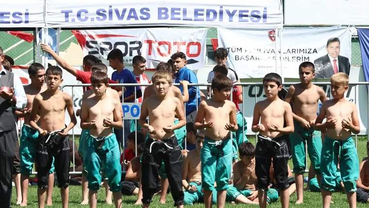Yeşil ve siyah güreş kıyafetleri giyen çocuklar çim alanda sıralanıyor; arkada T.C. Sivas Belediyesi pankartı var.