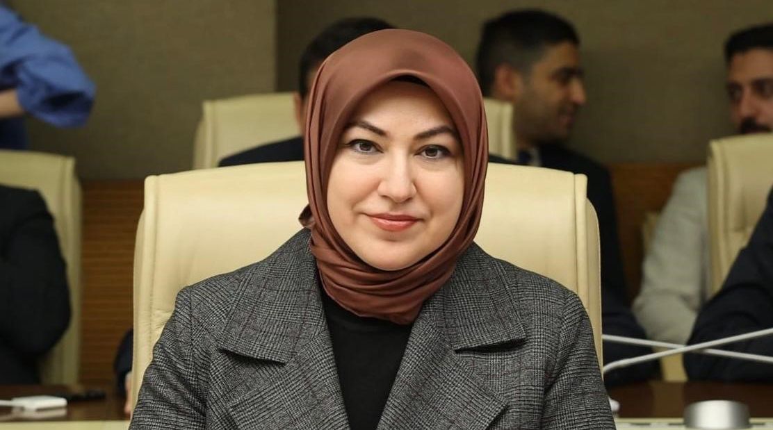 Toplantı salonunda başörtülü bir kadın, gri ceketle kameraya bakıyor; arka planda başka kişiler görülüyor.