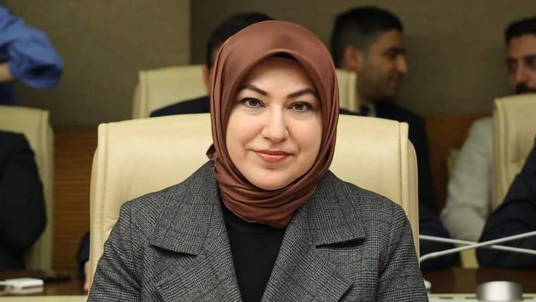 Toplantı salonunda başörtülü bir kadın, gri ceketle kameraya bakıyor; arka planda başka kişiler görülüyor.