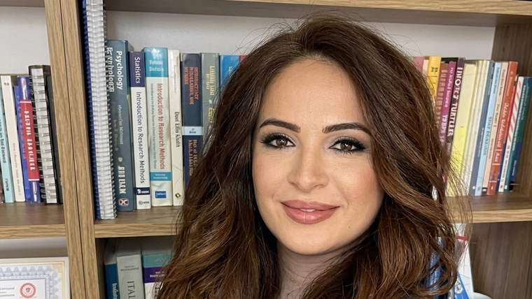 Kitaplık önünde poz veren, uzun kahverengi saçlı bir kadın; raflarda kitaplar ve çerçeveli belge görülüyor.