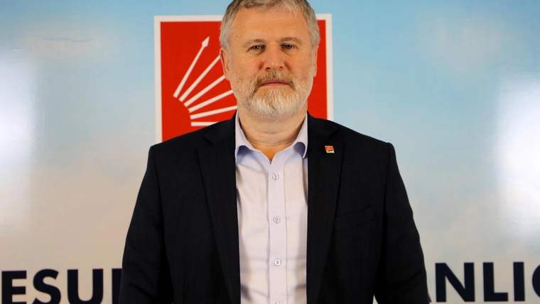 Sakallı bir erkek, kırmızı-beyaz amblemli ve yazılı arka plan önünde siyah ceketle kameraya bakıyor.