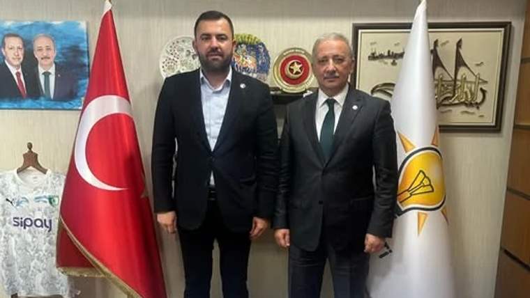 Ofis ortamında iki erkek, Türk bayrağı ve üzerinde amblem bulunan beyaz bayrağın önünde yan yana duruyor.