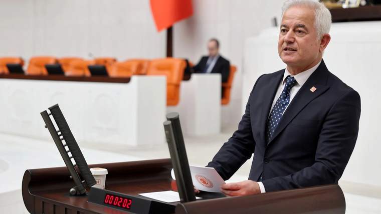 Kürsüde duran beyaz saçlı bir kişi, elindeki kâğıda bakarken meclis salonunda konuşuyor.