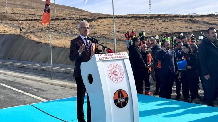 Bir kişi, açılış töreninde mikrofonla konuşuyor; arka planda inşaat işçileri ve bayraklar var.