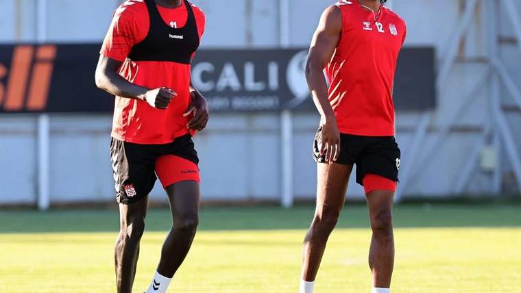 Çim sahada kırmızı antrenman formaları ve siyah şortlar giyen iki futbolcu yan yana koşuyor.