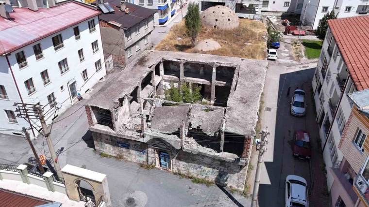 Kent içindeki yıkık iki katlı yapının çatısı ve duvarları kısmen çökmüş; çevresinde binalar ve araçlar görülüyor.