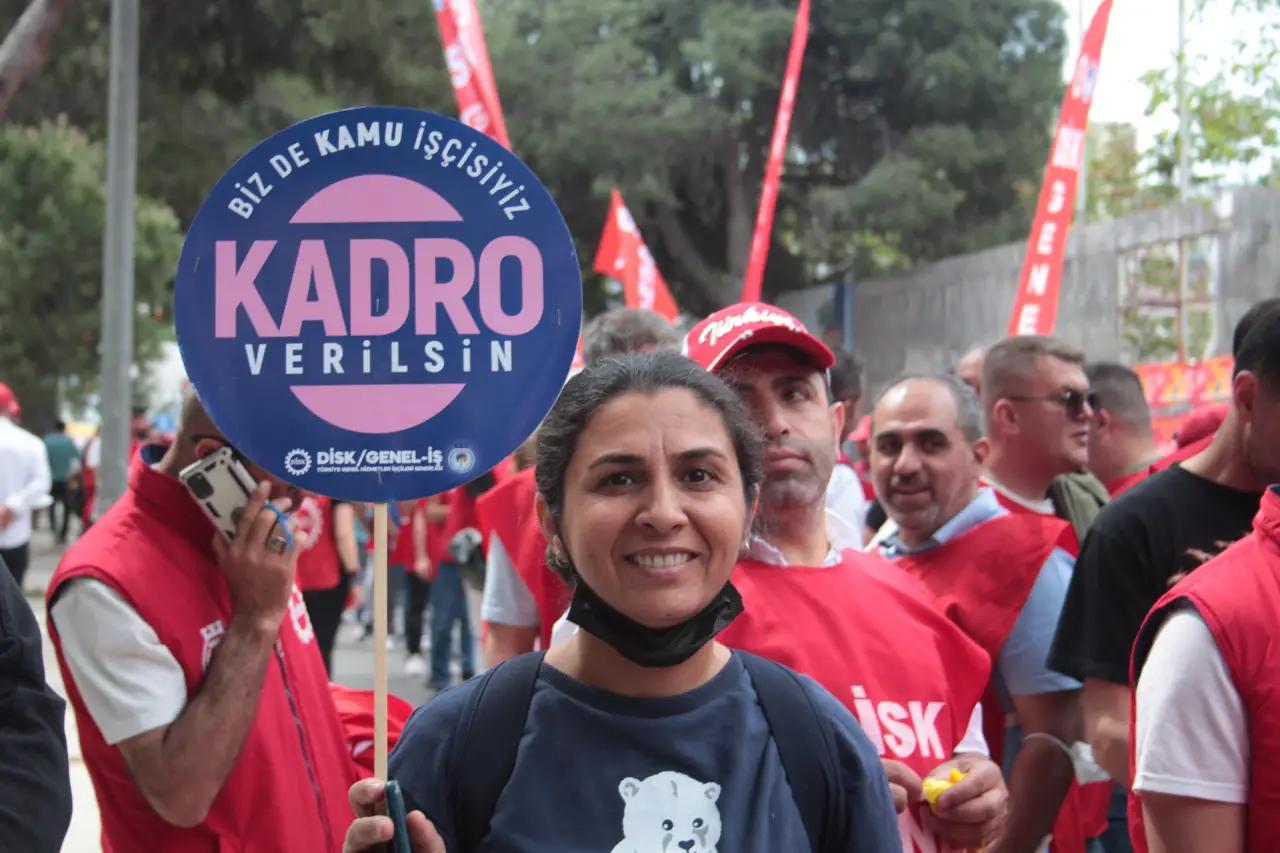 Önde maskesi çenesinde bir kadın, arkasındaki kırmızı tişörtlü kalabalıkla birlikte “Kadro verilsin” yazılı döviz taşıyor.