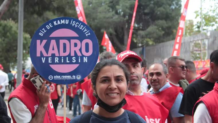 Önde maskesi çenesinde bir kadın, arkasındaki kırmızı tişörtlü kalabalıkla birlikte “Kadro verilsin” yazılı döviz taşıyor.