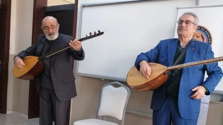 İki kişi, sınıf ortamında bağlama çalarken görülüyor; arkalarında beyaz tahta ve boş sandalye bulunuyor.