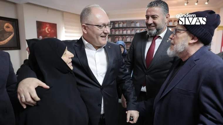 Kapalı bir mekânda dört kişi sohbet ediyor; ortadaki erkek, siyah başörtülü kadının omzuna elini koyuyor.