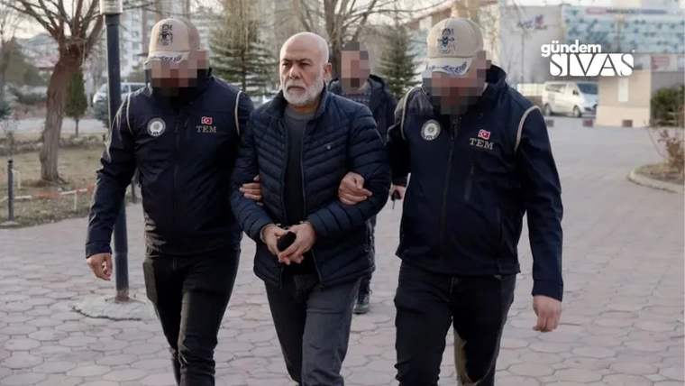 Ortadaki sakallı bir erkek, iki üniformalı görevlinin kolları arasında kaldırımda yürürken görülüyor.
