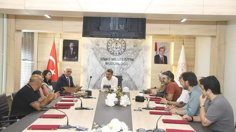 Toplantı salonunda uzun masa çevresinde oturan kişiler, ortadaki konuşmacıyı dinliyor; duvarda kurum yazısı bulunuyor.