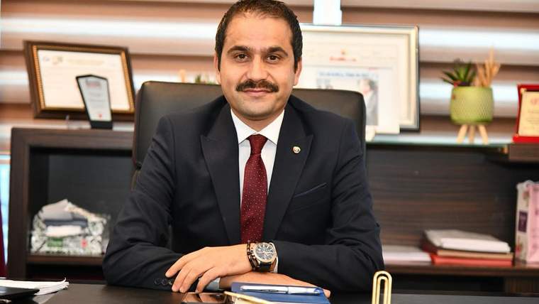 Masa başında oturan bıyıklı bir adam, koyu renk takım elbisesiyle kameraya bakıyor; arka planda belgeler var.