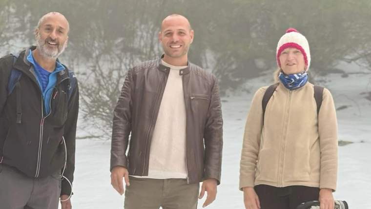 Karlı ve ağaçlık bir alanda üç kişi ayakta duruyor; sağdaki kişi elinde siyah bir ekipman taşıyor.