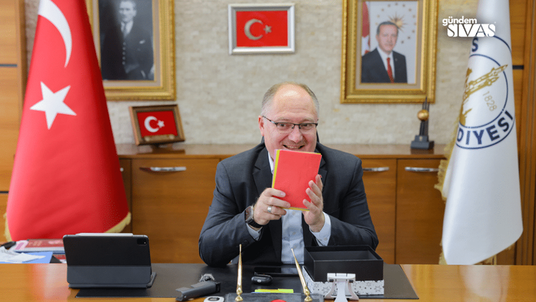 Bir masa etrafında oturan bir kişi, elinde kırmızı bir defter tutuyor ve gülümsüyor.