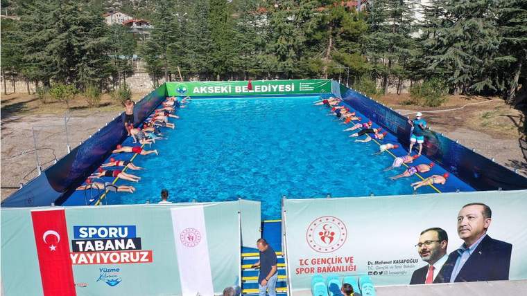 Üzerinde Akseki Belediyesi yazan portatif havuzda çocuklar su içinde, çevresinde spor temalı pankartlar görülüyor.