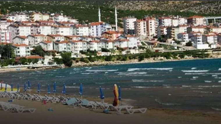 Kıyıda kapalı renkli şemsiyeler ve boş şezlonglar, karşıda su kenarındaki yerleşim ve ağaçlı yamaç görülüyor.