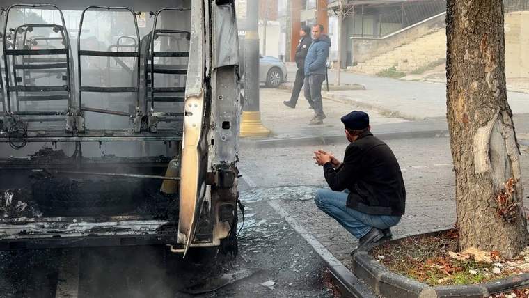 Yanmış bir servis aracının arka bölümü ve yanında çömelerek inceleyen bir kişi görülüyor.