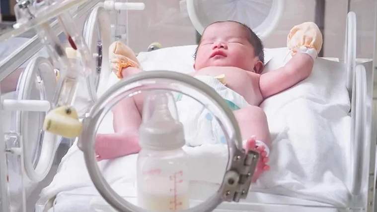 Yeni doğmuş bir bebek, hastane yatağında yatıyor, yanında bir bebek biberonu var.
