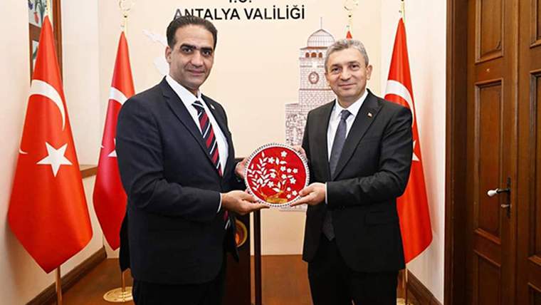 İki kişi, bir odada birbirlerine hediye takdim ediyor. Arka planda Türkiye bayrağı ve Antalya Valiliği tabelası var.