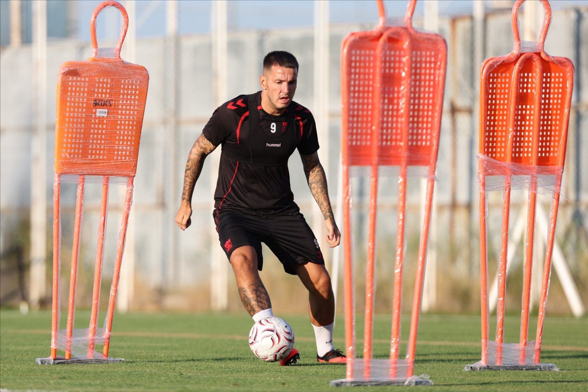 Siyah antrenman kıyafetli bir futbolcu, çim sahada top sürerken turuncu mankenlerin arasından geçiyor.