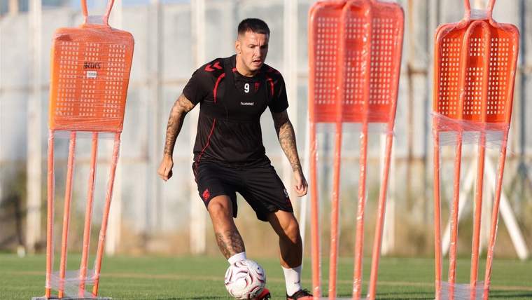 Siyah antrenman kıyafetli bir futbolcu, çim sahada top sürerken turuncu mankenlerin arasından geçiyor.