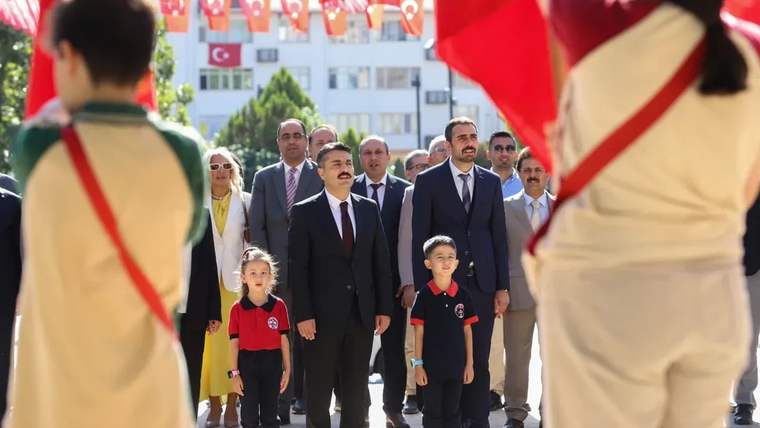 Türk bayraklarıyla süslenmiş alanda, iki izci çocuğun arasında yürüyen yetişkinler ve öğrenciler görülüyor.