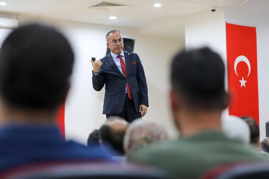 Kırmızı-beyaz Türk bayrağının asılı olduğu bir salonda, konuşma yapan bir kişi dinleyicilere hitap ediyor.