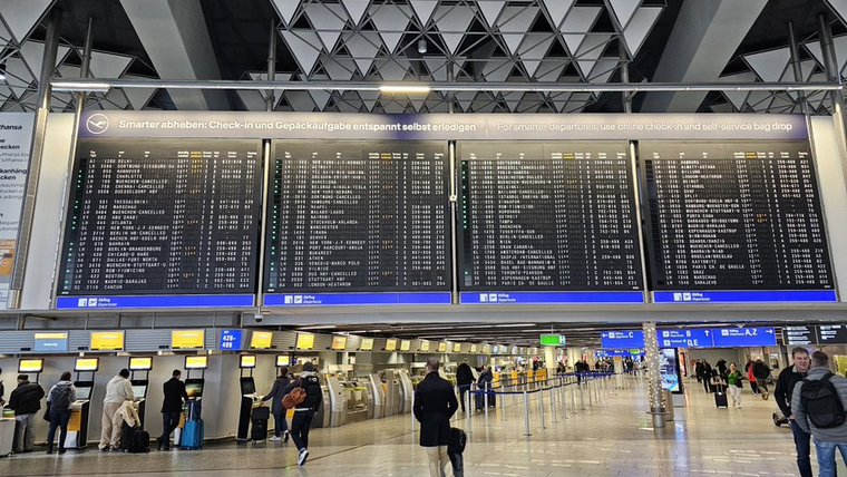Havalimanı terminalinde uçuş bilgi ekranları, check-in kontuarları ve bagajlarıyla bekleyen yolcular görülüyor.