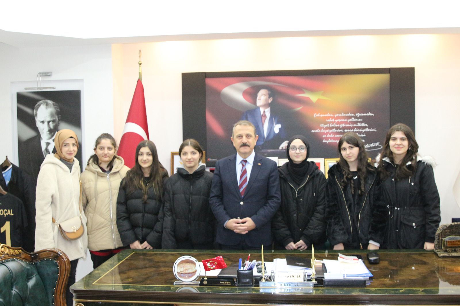 Bir odada, masanın arkasında duran bir erkek ve yanında sıralanan öğrencilerle Türk bayrağı görülüyor.