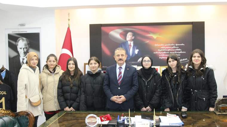 Bir odada, masanın arkasında duran bir erkek ve yanında sıralanan öğrencilerle Türk bayrağı görülüyor.