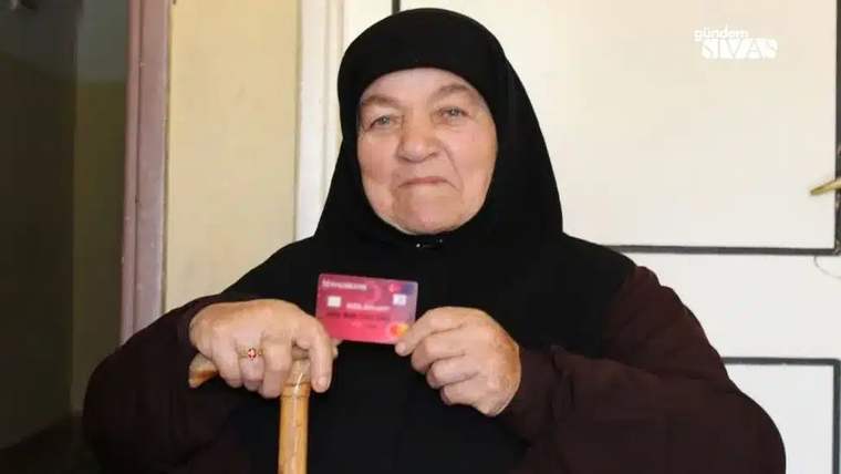 Başörtülü yaşlı bir kadın, elinde kırmızı bir kart ve bastonuyla kapalı bir mekânda duruyor.