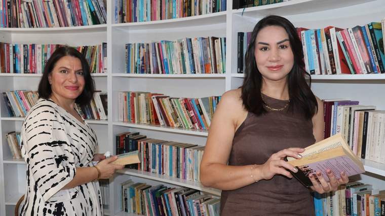 İki kadın, kitap raflarının önünde kitap tutarak poz veriyor.