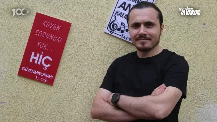 Duvar önünde kollarını bağlayan sakallı bir erkek, yanında “Güven sorunum yok... hiç” yazılı kırmızı levha.