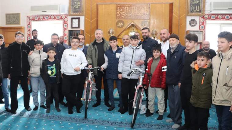 Cami içinde çocuklar ve yetişkinlerden oluşan bir grup, iki bisikletin yanında yan yana duruyor.