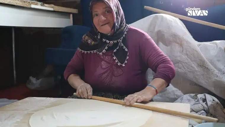 Başörtülü bir kadın, iç mekânda masanın üzerindeki hamuru uzun bir oklavayla açıyor.
