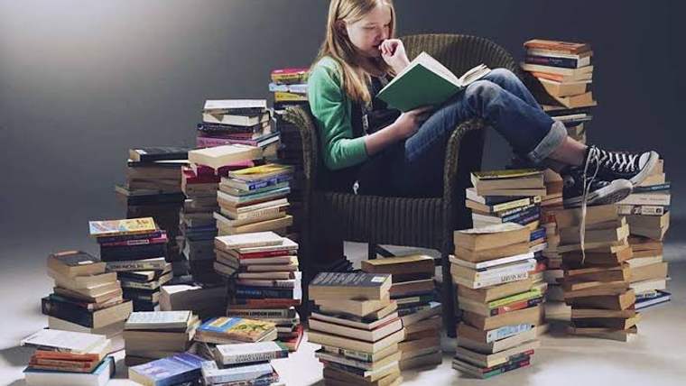 Bir koltukta oturan genç kız, etrafında kitap yığınlarıyla bir kitap okuyor.