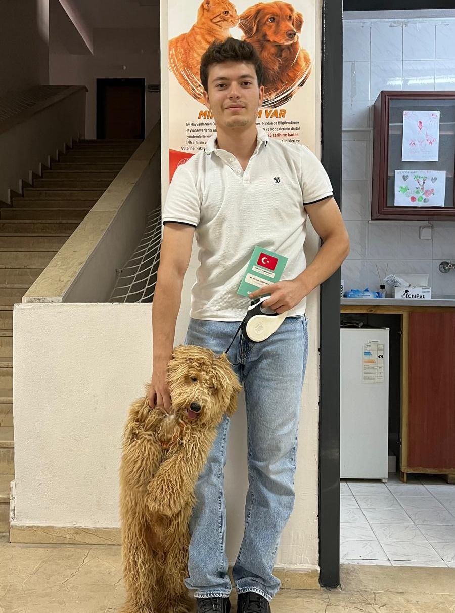 Bir kişi, tasma taktığı küçük tüylü köpeğin yanında dururken elinde Türk bayraklı bir kart tutuyor.
