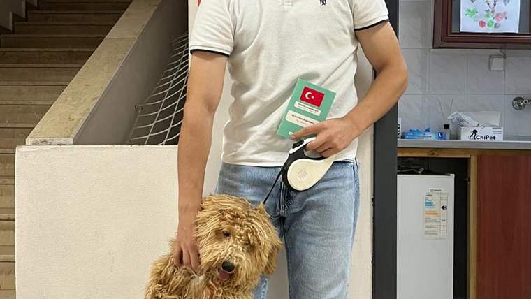 Bir kişi, tasma taktığı küçük tüylü köpeğin yanında dururken elinde Türk bayraklı bir kart tutuyor.